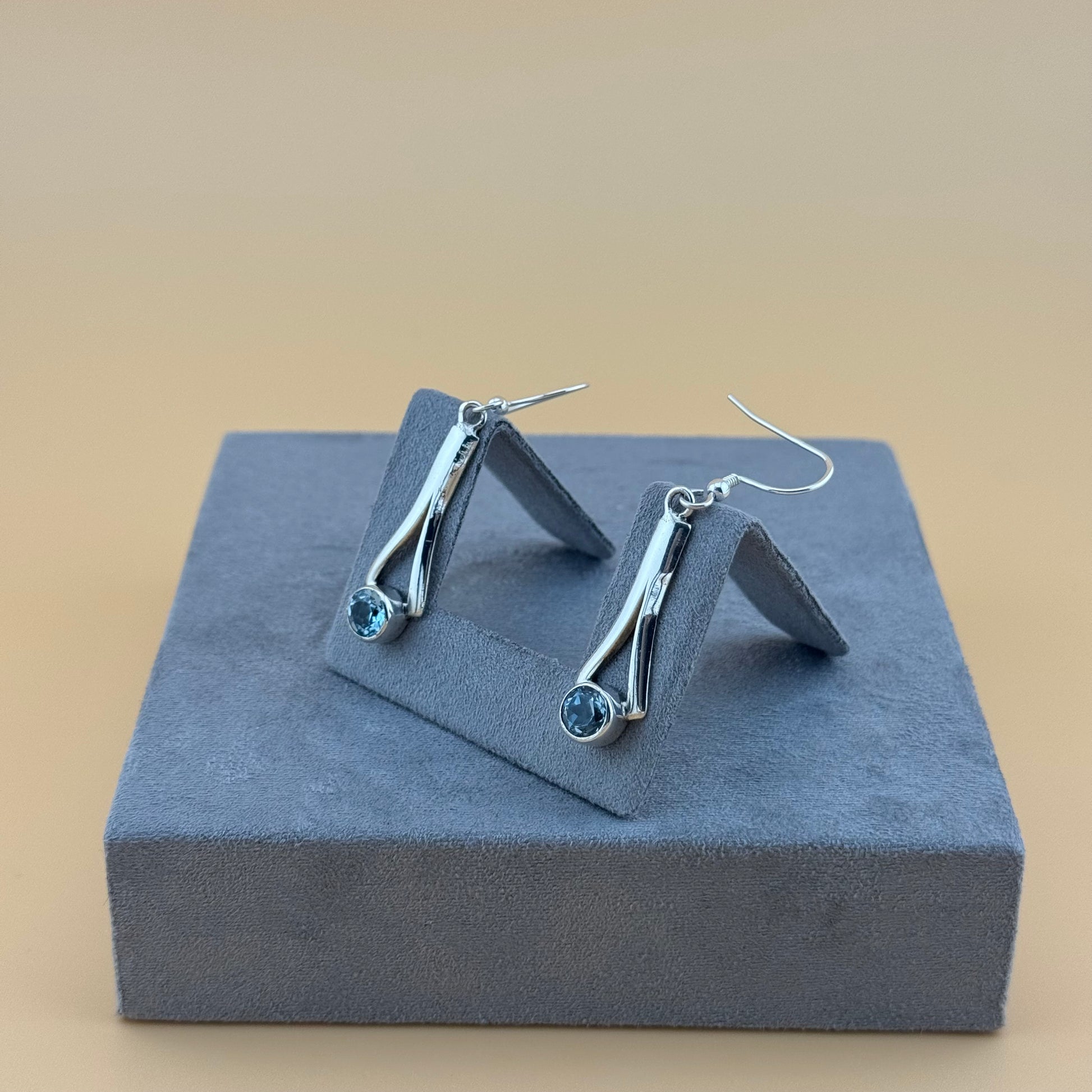 Pair of silver earrings with blue aquamarine gemstones on a gray stand against a beige background