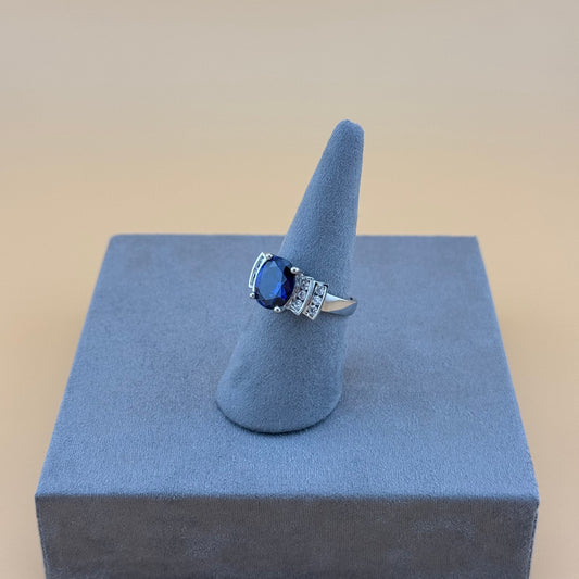 Silver ring with a blue sapphire gemstone on a gray stand against a beige background