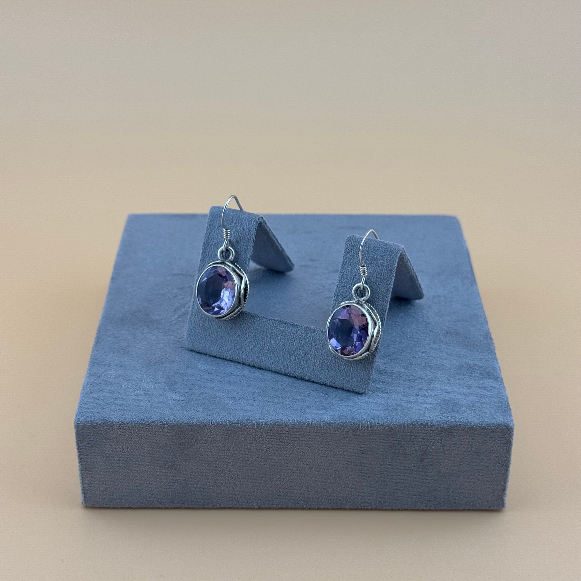 Pair of silver earrings with purple amethyst gemstones on a blue stand against a beige background