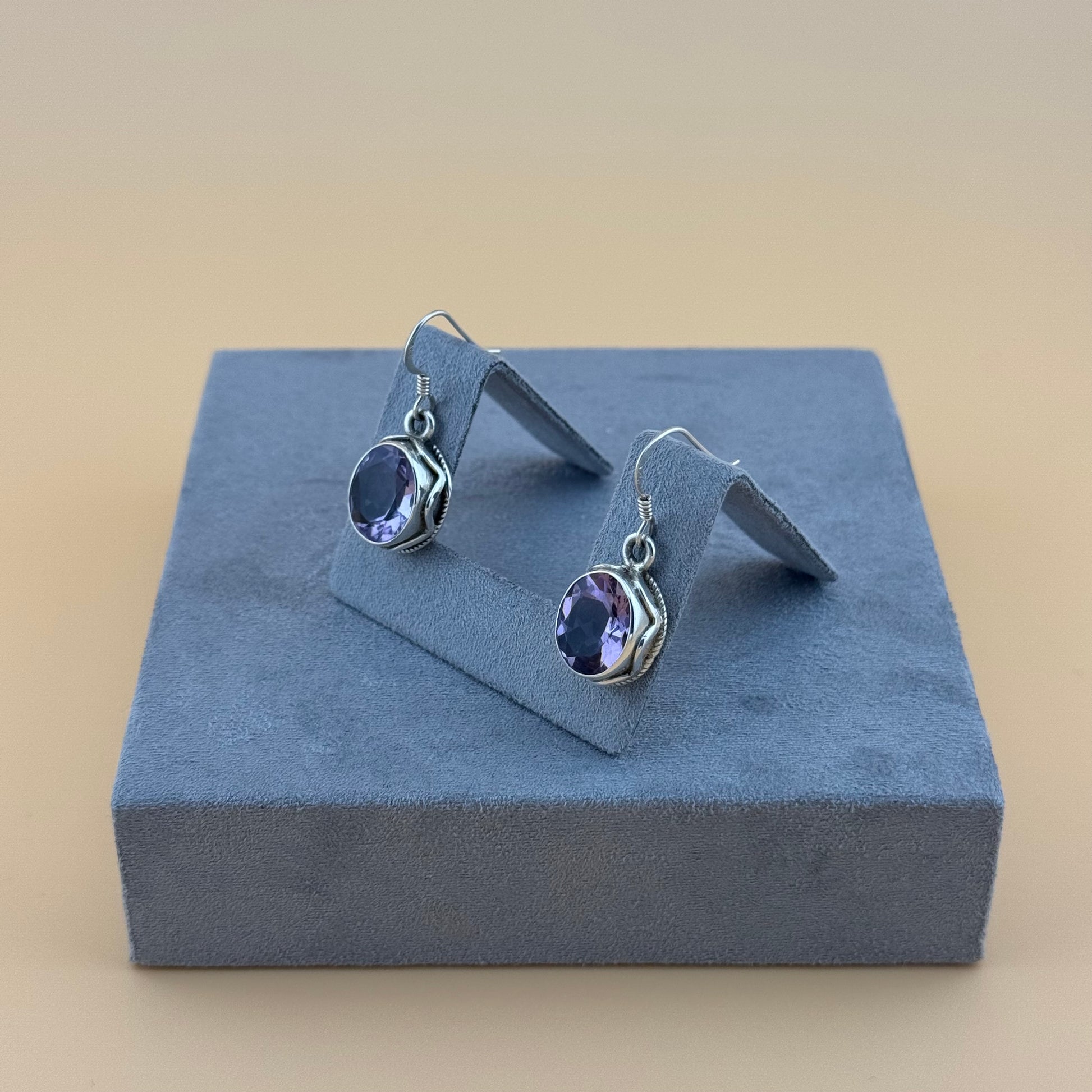Pair of silver earrings with purple amethyst gemstones on a blue stand against a beige background