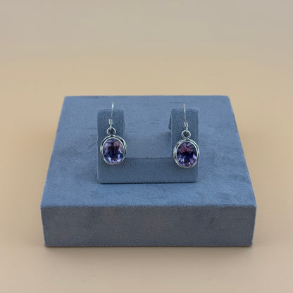 Pair of purple amethyst gemstone earrings on a blue stand against a beige background