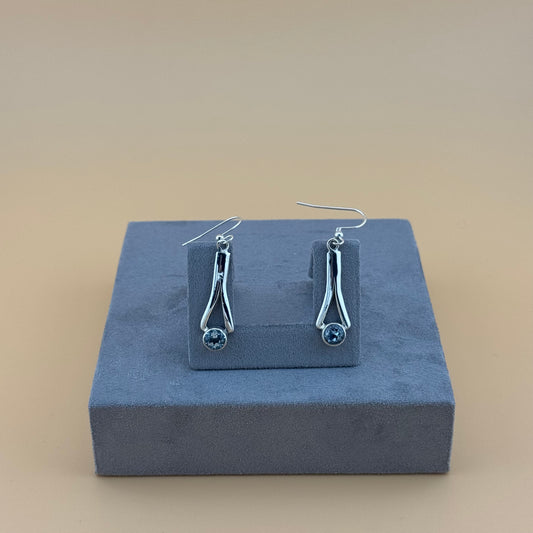 Silver earrings with blue aquamarine stones on a gray stand against a beige background
