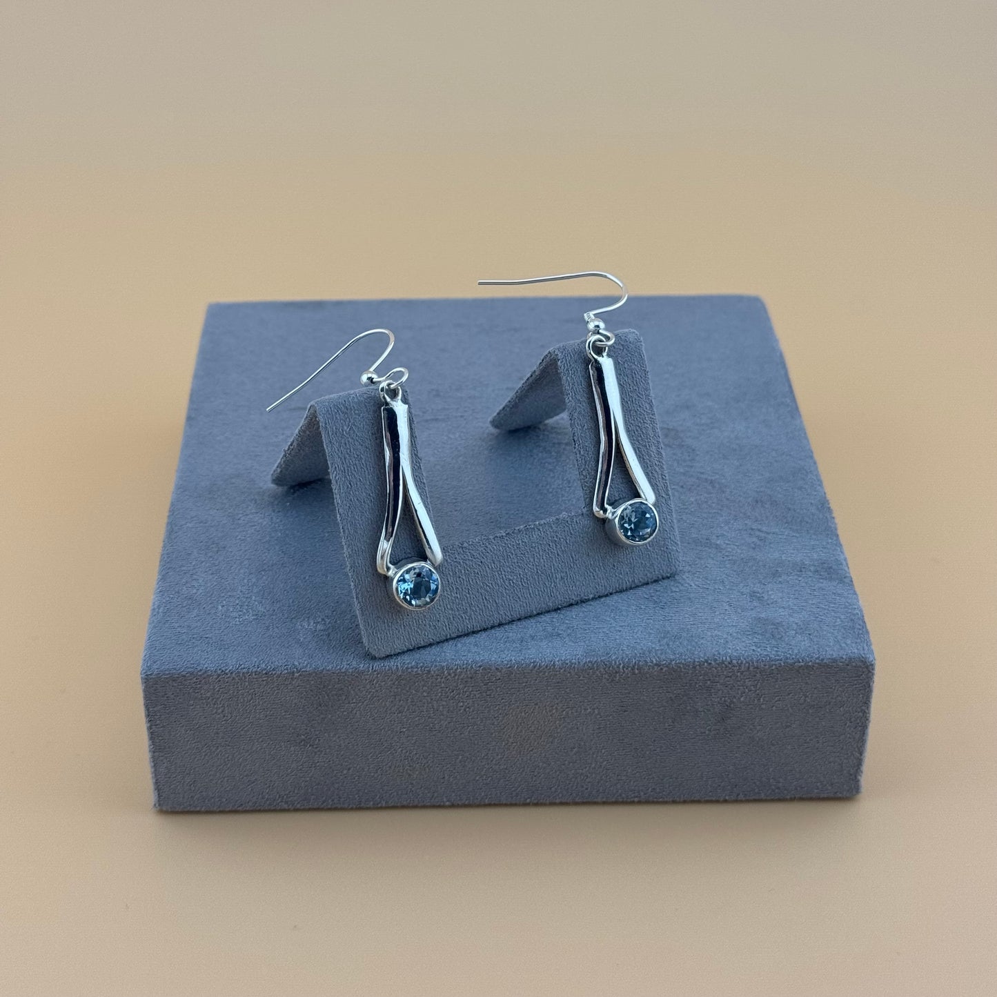 Silver earrings with blue aquamarine gemstones on a gray jewelry display box against a beige background