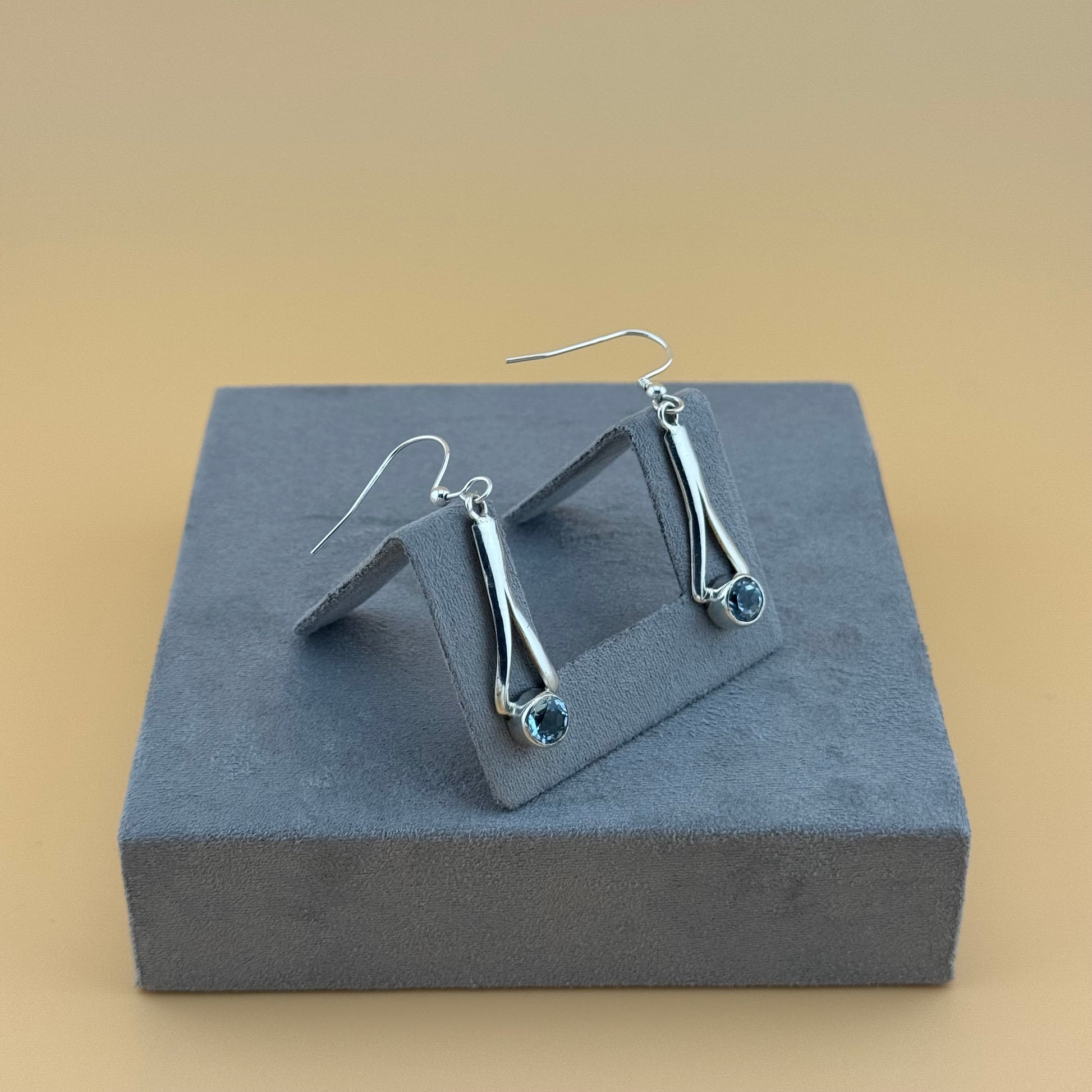 Silver earrings with blue aquamarine gemstones on a gray stand against a beige background